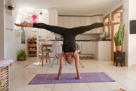 Téléchargez les photos : Corps complet d'athlète féminine en forme en vêtements de sport prenant la pose Adho Mukha Vrikshasana et faisant des fentes sur le tapis violet tout en pratiquant le yoga dans la cuisine à la maison - en image libre de droit