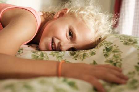 Foto de Vista lateral de la chica rubia sonriente acostada en la cama y mirando a la cámara mientras se relaja en el dormitorio - Imagen libre de derechos