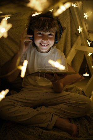 Foto de Cuerpo completo de chico positivo con auriculares viendo vídeo en la tableta y mirando a la cámara mientras está sentado cerca de lámparas brillantes en la habitación acogedora - Imagen libre de derechos