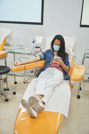 Photo for From above full length of adult woman in casual outfit and medical mask donating blood in modern laboratory while texting message on smartphone - Royalty Free Image