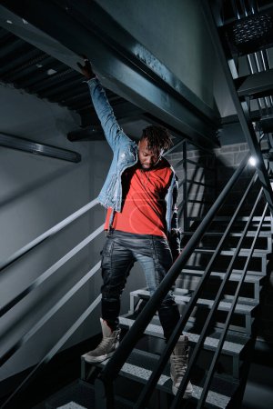 Photo for Full body of black man standing on metal stairs in dark building and looking down while putting on denim jacket - Royalty Free Image