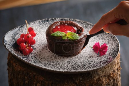 Photo for From above anonymous person using spoon to take piece of chocolate fondant with red currant jam and mint on plate with powdered sugar - Royalty Free Image