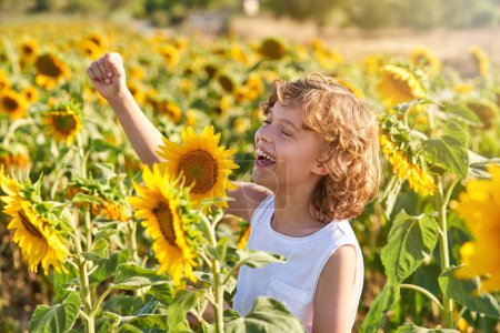 Photo for Happy child with blond curly hair laughing while looking at blooming sunflowers in field on sunny summer day - Royalty Free Image