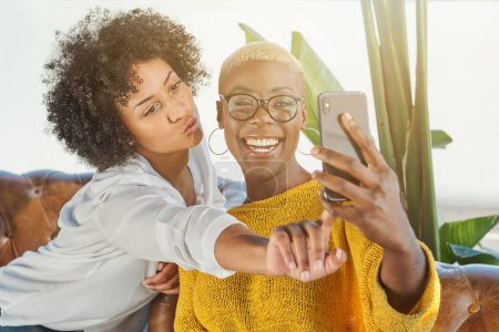 Photo for Positive multiethnic female friends laughing and making duck face while taking self portrait on smartphone and sitting on leather couch together - Royalty Free Image