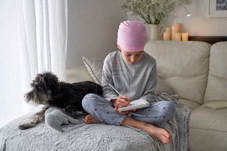 Photo for Full body of focused barefoot boy in pink bandana as symbol of breast cancer campaign and awareness writing in notebook near Miniature Schnauzer dog on sofa - Royalty Free Image
