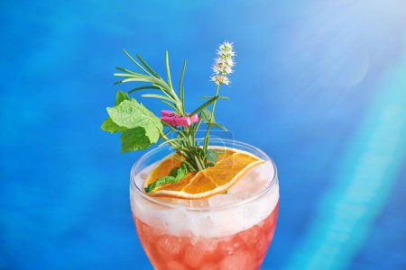 Photo for From above glass of cold refreshing cocktail drink decorated with orange slice and peppermint rosemary leaves in ice cubes while placed on blue background - Royalty Free Image