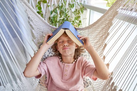 Foto de Niño con los ojos cerrados descansando en hamaca de punto con libro en la cabeza y pensando en la historia en el balcón en casa en brillante día de fin de semana - Imagen libre de derechos