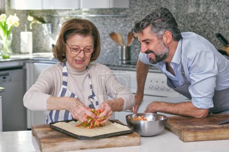 Téléchargez les photos : Homme positif dans le tablier regardant la mère étaler le remplissage sur la pâte non cuite tout en se préparant pour la cuisson de tarte dans la cuisine légère - en image libre de droit