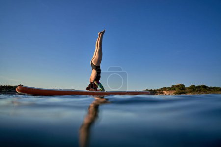 Téléchargez les photos : Vue latérale pleine longueur de femme flexible en maillot de bain debout sur le paddleboard à Salamba Shirshasana tout en pratiquant le yoga en mer le soir - en image libre de droit