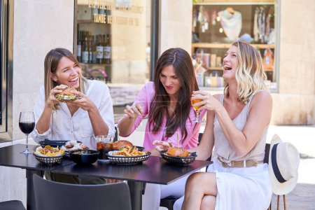Téléchargez les photos : Rire de jeunes femmes aux cheveux lâches assis à table sur la terrasse du café tout en dégustant des hamburgers délicieux avec des frites et divers alcools et en s'amusant - en image libre de droit