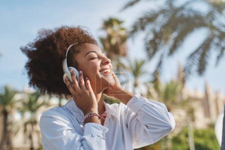 Foto de Joven mujer afroamericana alegre en auriculares sonriendo y escuchando música en el parque verde de la ciudad en el día de verano - Imagen libre de derechos