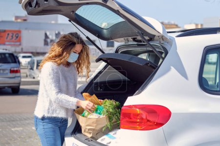 Téléchargez les photos : Calme jeune femme en pull blanc et masque protecteur vérifier les achats d'épicerie dans des sacs à provisions placés dans le coffre de la voiture sur le parking ensoleillé du supermarché - en image libre de droit