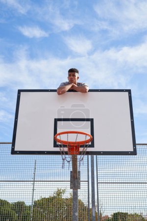 Photo for From below of basketball player looking away while resting on backboard placed on sports ground against blue sky - Royalty Free Image