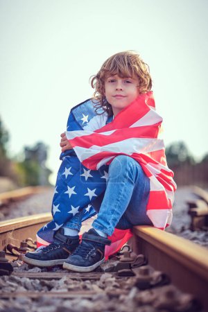 Téléchargez les photos : Contenu enfant aux cheveux blonds regardant la caméra assis sur le chemin de fer pendant la fête de l'indépendance américaine sur fond flou - en image libre de droit
