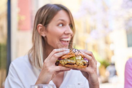 Téléchargez les photos : Jeune femme positive avec les cheveux lâches en chemisier blanc appréciant hamburger succulent appétissant tout en déjeunant au café extérieur - en image libre de droit