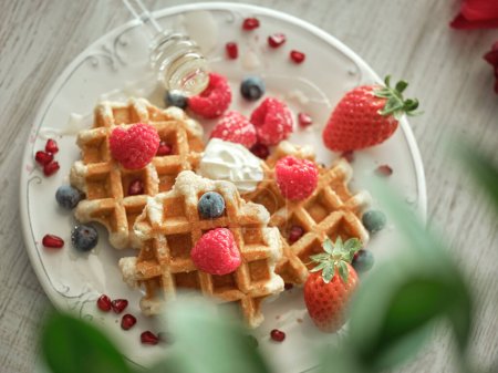 Photo for From above of sweet waffles served on plate with ripe berries and honey dipper in light kitchen with blurred green plant - Royalty Free Image