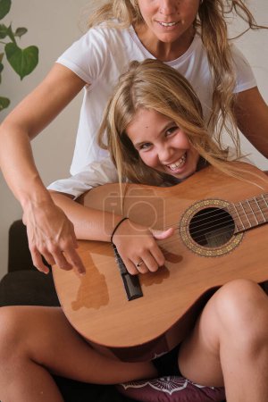 Foto de Emocionada joven mujer positiva con sonrisa dentada sentada en el sofá y tocando la guitarra de madera acústica mientras mira a la cámara - Imagen libre de derechos