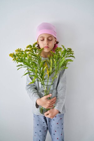 Photo for Tranquil cute child with closed eyes wearing pink headband while standing on white backdrop with vase of blooming Solidago - Royalty Free Image