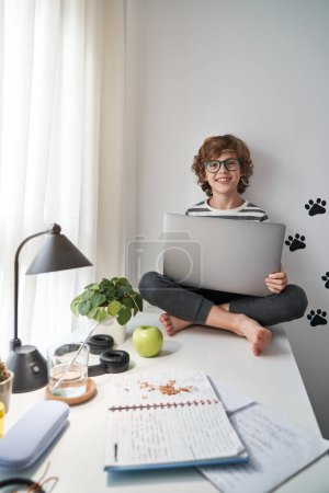 Photo for Full body of positive kid in glasses looking at camera and browsing laptop while doing homework in light bedroom near window - Royalty Free Image