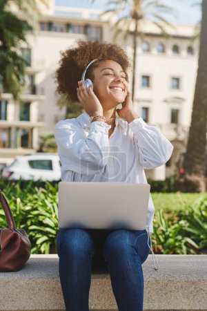 Foto de Mujer afroamericana sonriente con auriculares trabajando remotamente en el portátil y escuchando música mientras está sentada en el banco de piedra en el parque verde de la ciudad - Imagen libre de derechos
