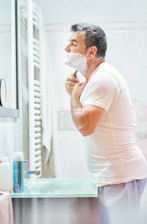 Foto de Vista lateral de la cara de afeitado masculino de pelo gris maduro con afeitadora por encima del fregadero mientras se mira en el espejo - Imagen libre de derechos