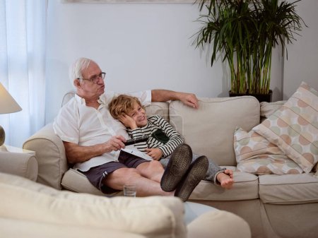 Foto de Cuerpo completo de nieto encantado viendo películas con un hombre mayor mientras descansa en el sofá en la sala de estar ligera en casa - Imagen libre de derechos