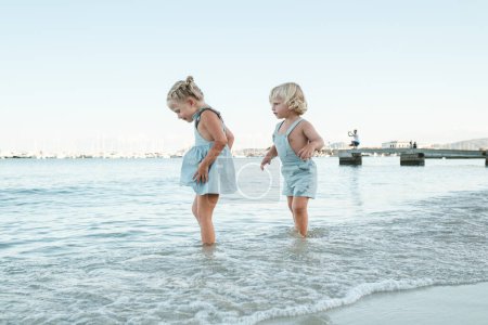 Téléchargez les photos : Vue latérale de mignons petits enfants marchant dans les eaux peu profondes de la mer tout en profitant des vacances d'été sur la plage - en image libre de droit