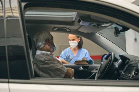 Téléchargez les photos : Cultivez le médecin féminin dans un masque respiratoire en parlant avec un patient âgé de sexe masculin en auto tout en se regardant pendant l'examen - en image libre de droit