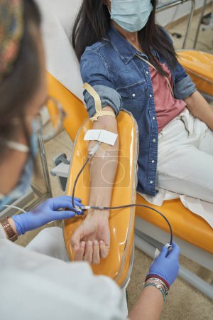 Foto de Enfermera de la cosecha en matorrales y guantes médicos que lleva a cabo el procedimiento de donación de sangre en la mujer en ropa casual y máscara protectora en el hospital - Imagen libre de derechos