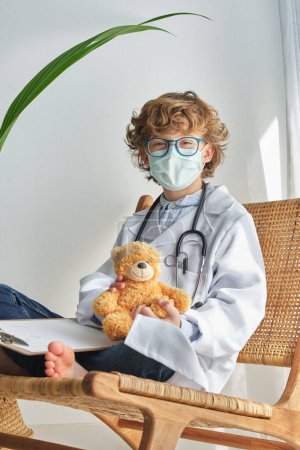 Foto de Niño en uniforme médico y gafas sentado con las piernas cruzadas y oso suave en la silla de mimbre mientras mira a la cámara - Imagen libre de derechos