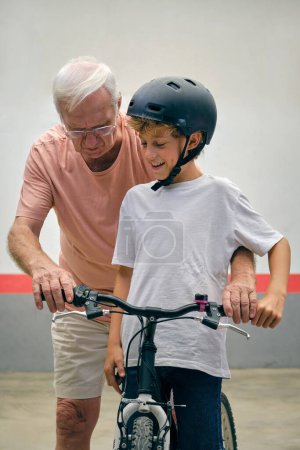 Téléchargez les photos : Homme âgé en tenue décontractée et lunettes aidant petit-fils sur vélo dans le casque tout en passant du temps ensemble contre le mur blanc - en image libre de droit