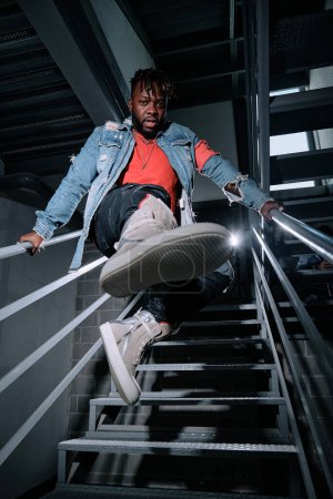 Photo for Full body of young ethnic male in trendy outfit and sneakers holding on railing without touching the stairs - Royalty Free Image