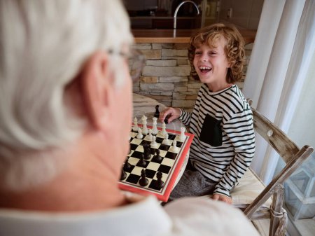 Téléchargez les photos : Petit-fils joyeux regardant la culture sans visage grand-père avec le sourire tout en jouant au jeu d'échecs ensemble à la table dans la cuisine légère à la maison - en image libre de droit