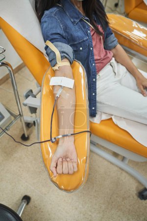Téléchargez les photos : De dessus de la culture femme anonyme en veste de denim assis dans le fauteuil et donnant du sang de la veine en laboratoire à l'hôpital - en image libre de droit