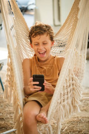 Téléchargez les photos : Ravi garçon pieds nus naviguant sur les médias sociaux sur téléphone portable et riant à la blague drôle tout en se relaxant dans l'hamac le week-end - en image libre de droit