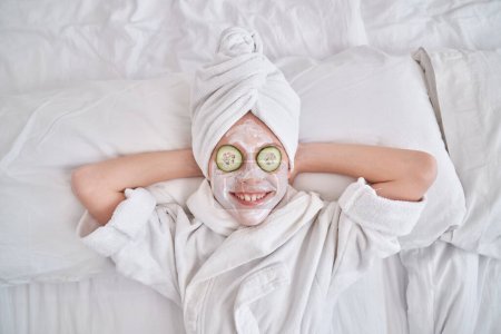Téléchargez les photos : Vue du dessus de l'enfant souriant avec masque à la crème faciale et tranches de concombre sur les yeux avec serviette sur la tête relaxant sur un lit confortable après la procédure de spa - en image libre de droit