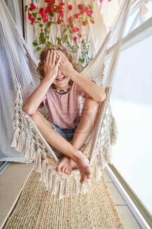 Photo for Full length of smiling barefoot kid in casual clothes sitting on hanging chair with closed legs and covering face with palms during sunny day - Royalty Free Image