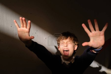 Photo for Excited preteen boy standing alone in dark room shouting and showing stop gesture with palms of hands while reacting and resisting sudden bright sunlight - Royalty Free Image