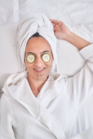 Photo for From above of relaxed female in bathrobe and towel with cucumber slices on eyes resting on bed during skincare procedure - Royalty Free Image