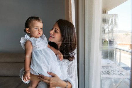 Téléchargez les photos : Curieux bébé en robe blanche regardant par la fenêtre avec intérêt dans les bras de la femme souriante dans le salon à la maison - en image libre de droit
