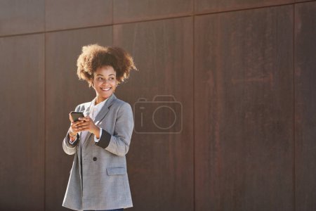 Foto de Mujer étnica feliz con el pelo rizado mirando hacia otro lado mientras está de pie cerca de la pared gris y el teléfono inteligente de navegación en el sol - Imagen libre de derechos