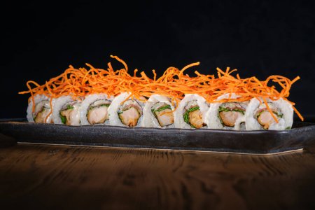 Téléchargez les photos : Rangée de sushi maki au saumon décoré de carotte râpée servie sur une assiette posée sur une table en bois sur fond noir - en image libre de droit