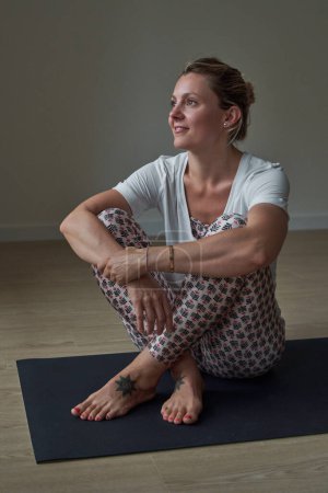 Foto de Cuerpo completo de hembra positiva en ropa deportiva sentada en una colchoneta de ejercicio y mirando hacia otro lado después de practicar yoga en el estudio - Imagen libre de derechos