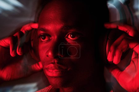 Foto de Varón afroamericano serio escuchando música en auriculares inalámbricos en la oscuridad con iluminación roja - Imagen libre de derechos