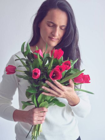 Téléchargez les photos : Portrait de femme calme en pull blanc debout avec bouquet de fleurs et les yeux fermés sur fond blanc - en image libre de droit
