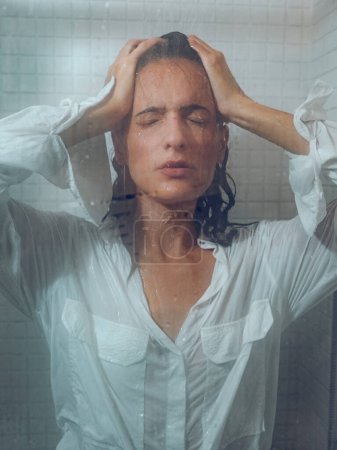 Téléchargez les photos : Femme sensuelle aux cheveux foncés et mouillés portant une chemise blanche debout sous l'eau dans la cabine de douche avec les yeux fermés - en image libre de droit