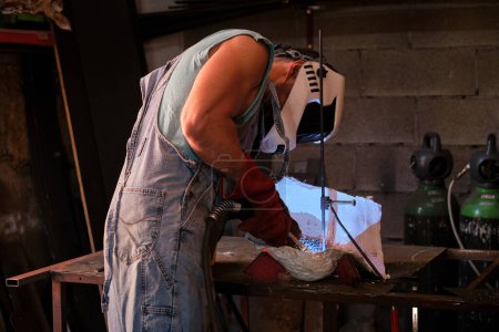 Foto de Vista lateral de un artesano masculino irreconocible en casco protector parado en el banco de trabajo y metal de soldadura en forma de yeso mientras trabaja en un taller profesional - Imagen libre de derechos