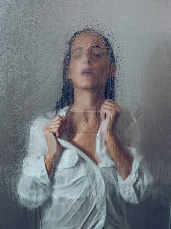 Foto de A través de vidrio de mujer sensible en blusa húmeda blanca de pie en cabina de ducha con gotas de agua y vapor en el baño - Imagen libre de derechos