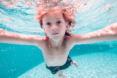 Téléchargez les photos : Vue sous-marine de l'enfant avec torse nu et en short de bain, enfant nageant dans de l'eau de piscine transparente propre et regardant la caméra - en image libre de droit