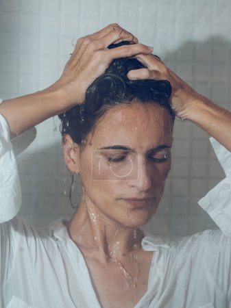 Foto de Hembra de pelo negro recogiendo el pelo mientras se cierran los ojos y conseguir completamente mojado en la ducha - Imagen libre de derechos